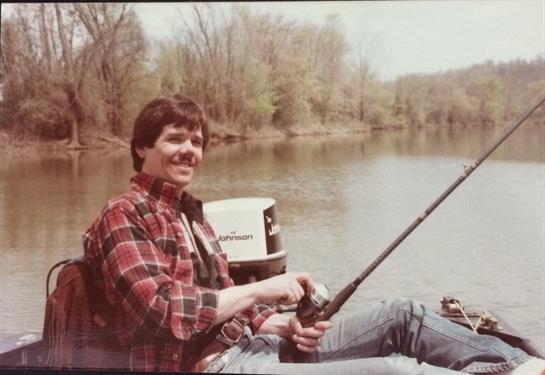 Man sits comfortably on a boat, fishing peacefully on a calm river surrounded by trees.