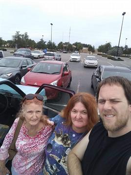 Three people pose together in a parking lot, sharing smiles under an overcast sky.