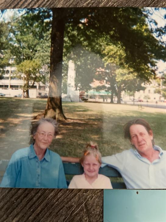 A woman, a child, and an elderly man sit on a bench in a park with trees around them.