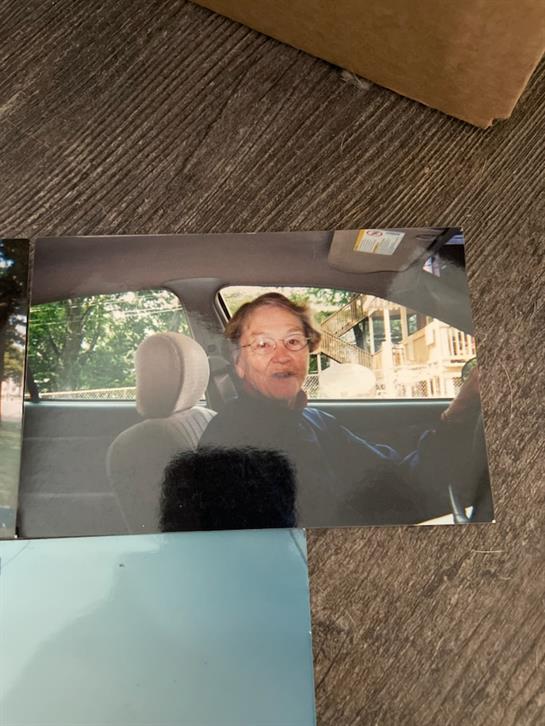 An elderly person is driving a car, looking surprised or amused. Trees surround the area.