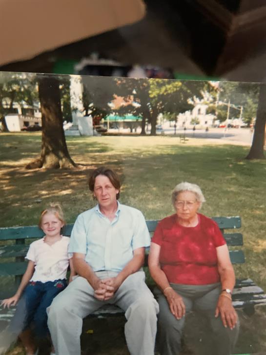 Three relatives relax on a bench in a park surrounded by trees and open space.