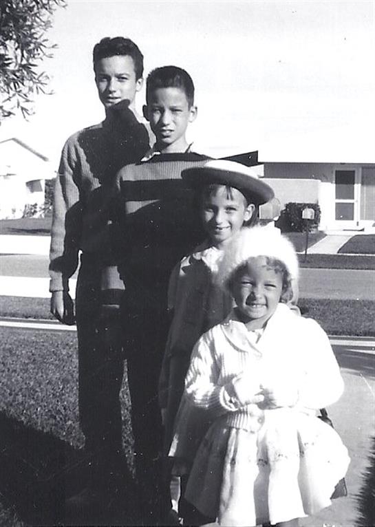 Four children stand together outside a house, smiling and dressed in vintage clothing.