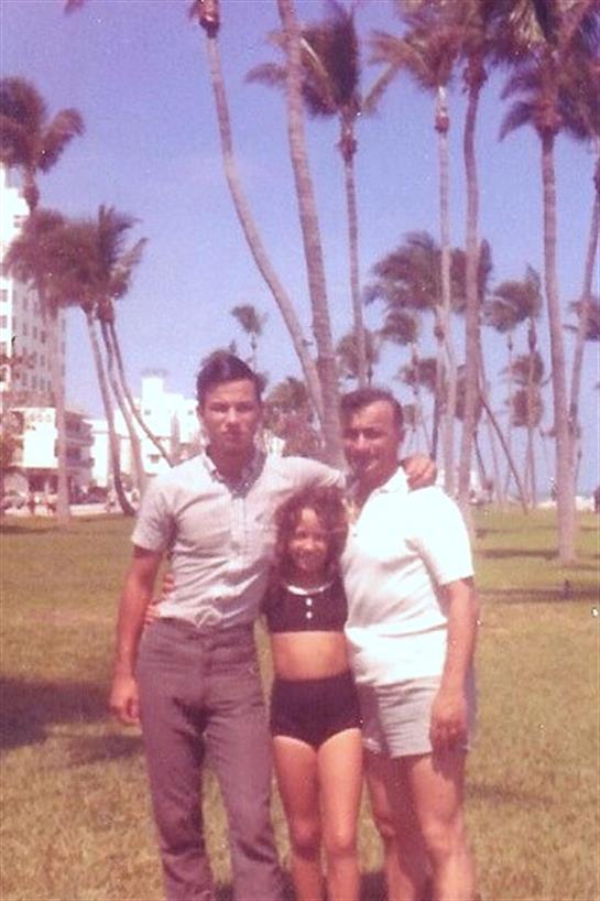 Two adults and a child pose happily in a park surrounded by palm trees on a sunny day.