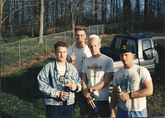 Four men stand together outside, holding drinks and smiling while enjoying a sunny day in nature.