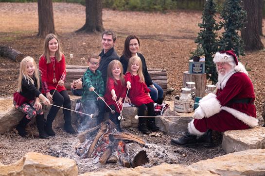 Children and adults sit by a warm fire, roasting marshmallows and enjoying festive cheer.