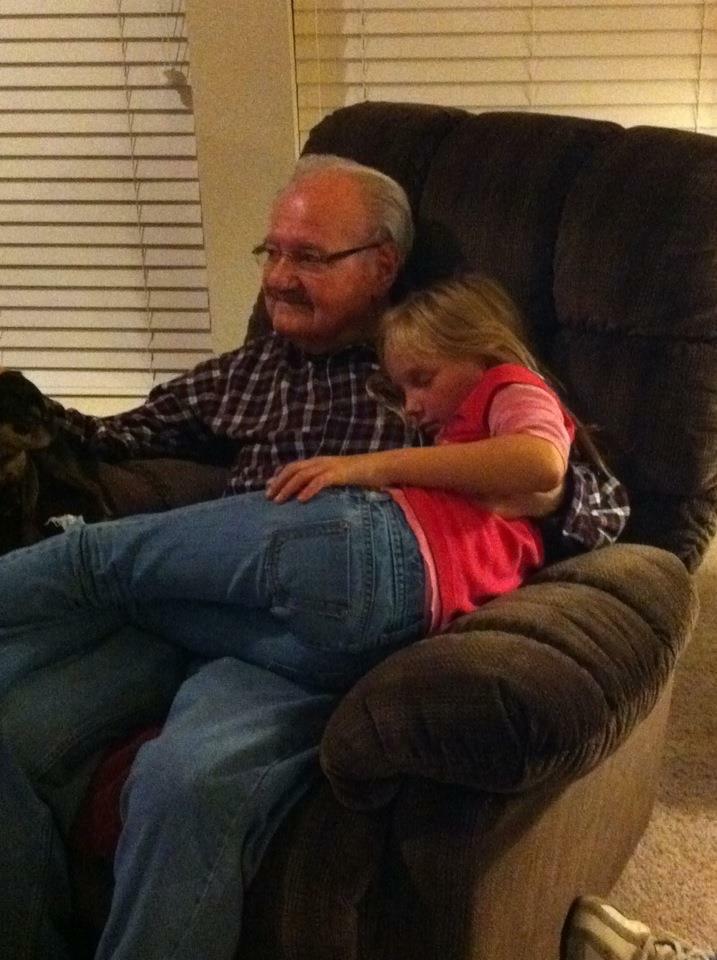 An elderly man sits comfortably with his granddaughter in an armchair during the evening.