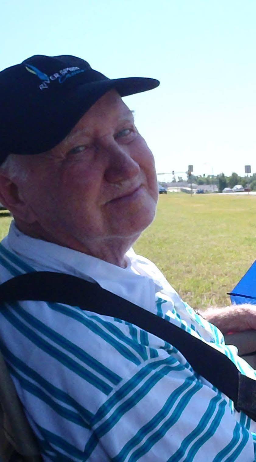 An elderly man sits contentedly outdoors, enjoying the warm sun and clear sky while smiling.