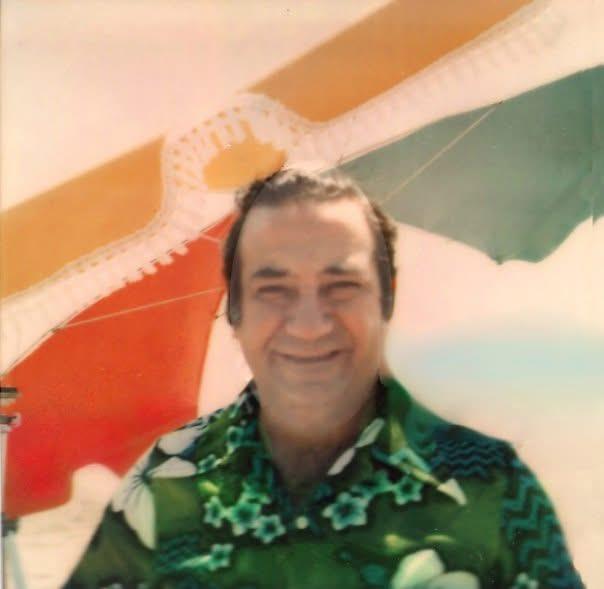 A cheerful man in festive attire stands by a vibrant banner at a summer gathering.