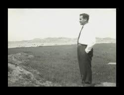 A man in formal attire gazes into the horizon while standing on a grassy hillside.
