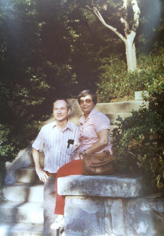 A cheerful older couple poses together in a vibrant garden filled with greenery and flowers.