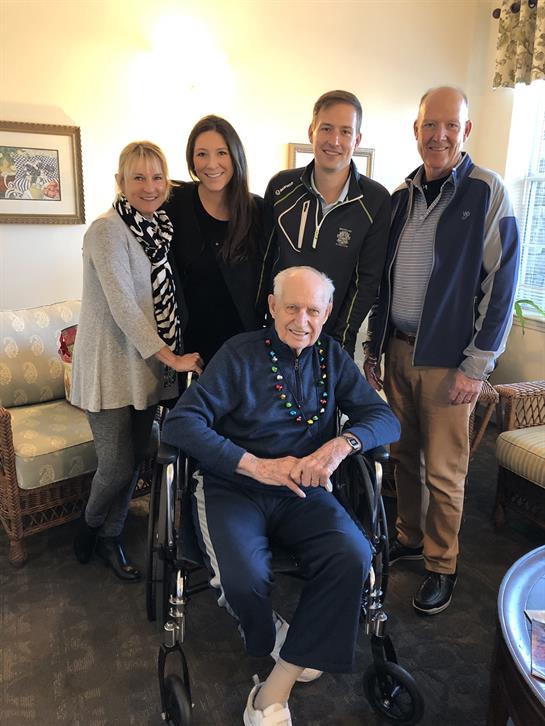 Family members pose happily with an elderly man in a wheelchair, enjoying a warm indoor atmosphere.