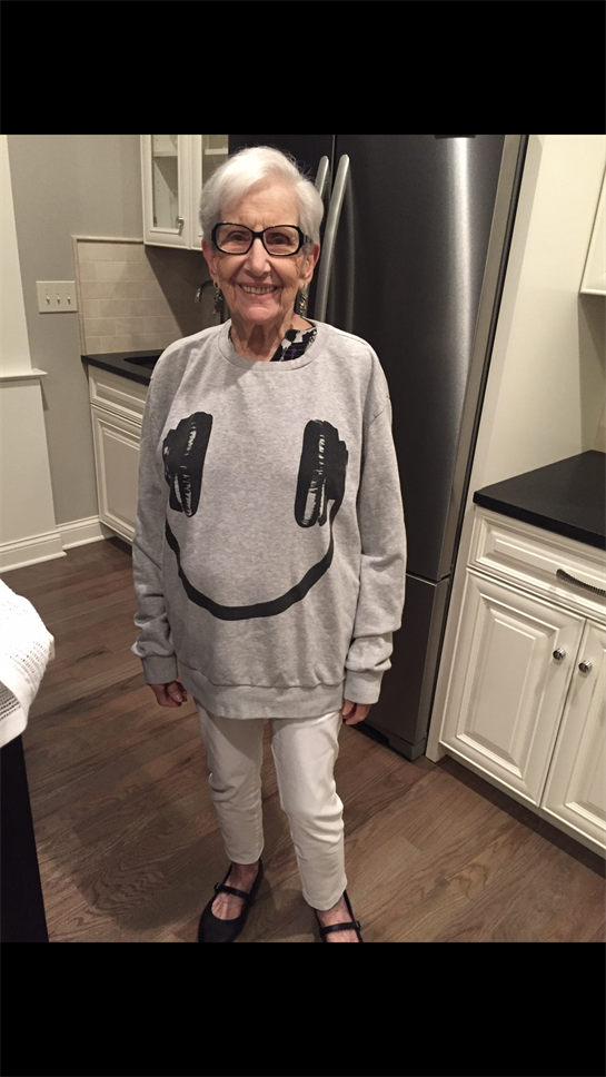 Older woman stands happily in her kitchen, showcasing a smiley face sweatshirt and casual attire.