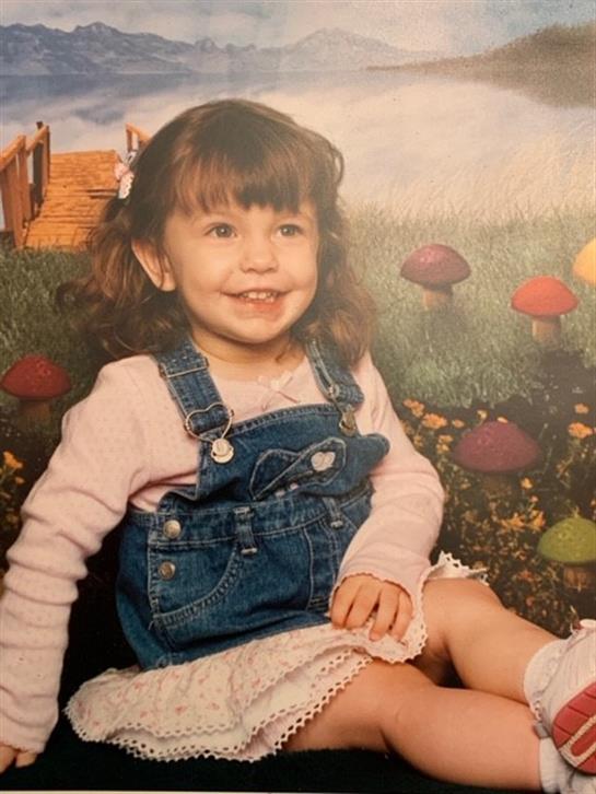 A cheerful little girl in denim overalls sits in a vibrant garden, surrounded by colorful mushrooms.