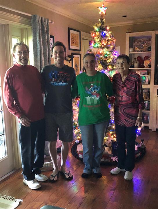 Four family members stand together beside a colorful Christmas tree in a cozy living room.