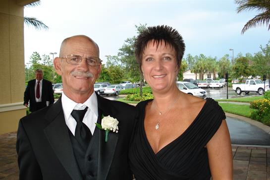 Older man in a suit smiles next to a woman in an elegant dress during a special occasion outdoors.