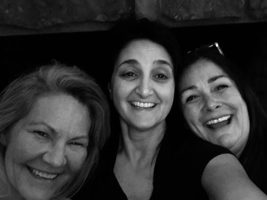 Three women share laughter and smiles while posing closely together during a joyful moment indoors.