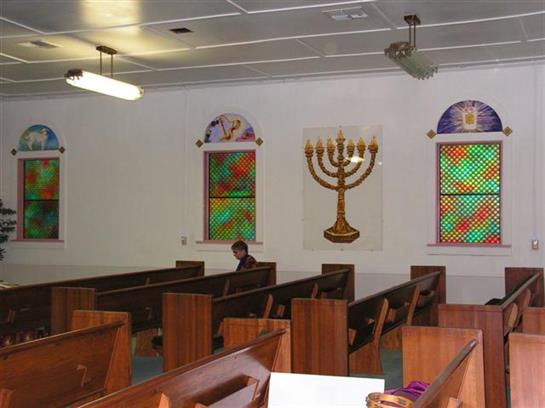 A person sits alone in a synagogue, surrounded by wooden benches and colorful stained glass.