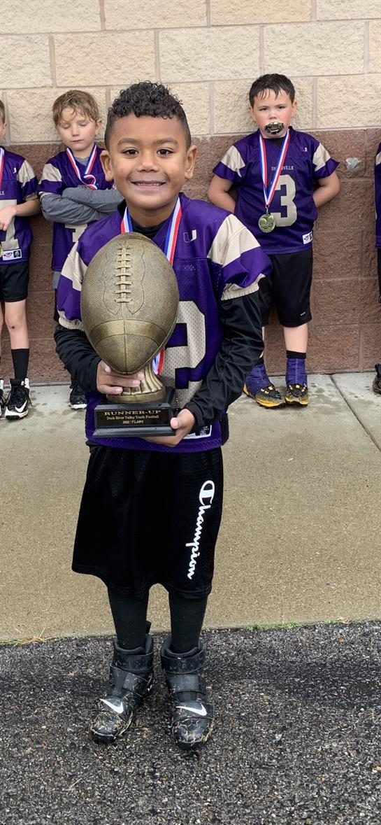 A young player beams with pride, holding a trophy with teammates after a football game.