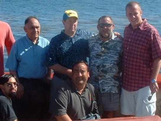 A group of five men stands together in front of a calm ocean, enjoying the sunny weather.