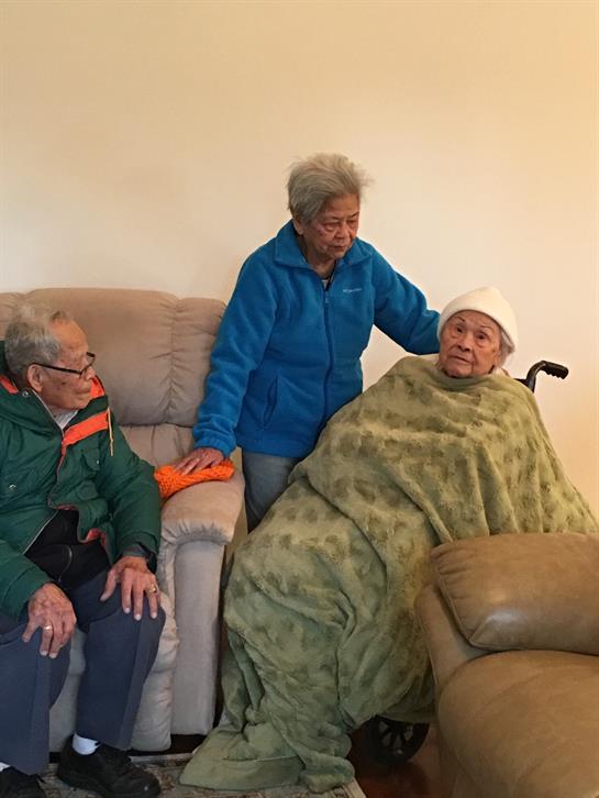 Three family members comfort an elderly woman in a warm living room, wrapped in a blanket.