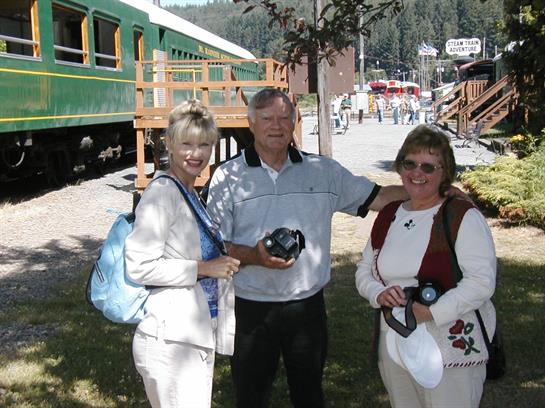 Three friends pose happily outside a mountain train station, capturing memories on a sunny day.