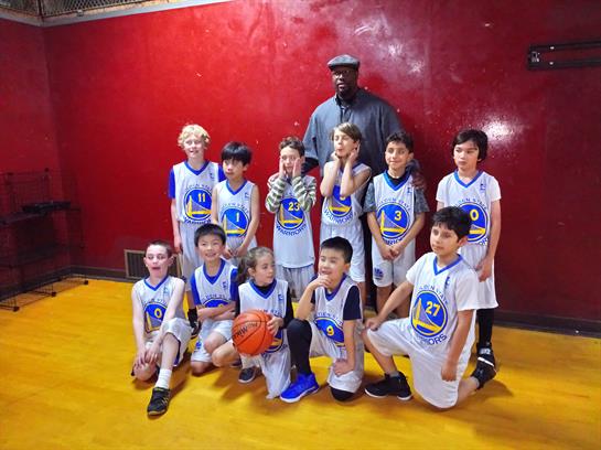 A youth basketball team celebrates their game while posing with a tall figure in a gym.