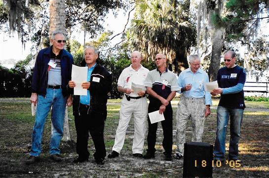 Men are gathered in a park, reading from papers, engaged in a community speech event outdoors.