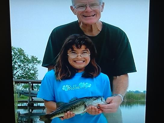Grandfather and granddaughter smile as they hold a large fish by the water's edge in summer.