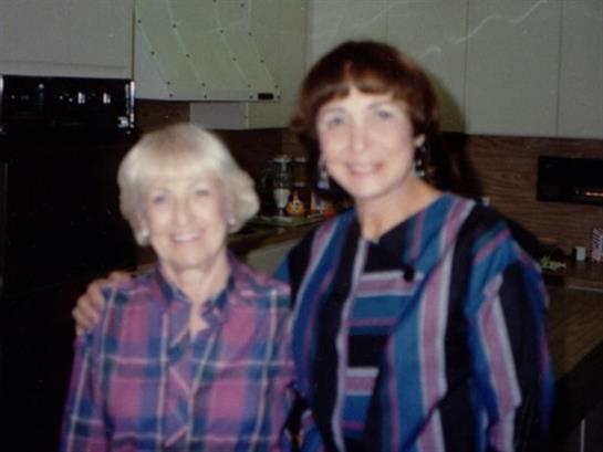 Two women pose closely, smiling, wearing colorful clothing, in a warm gathering environment at home.