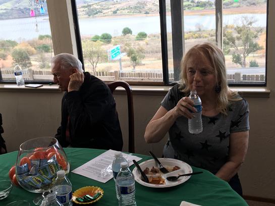 Two attendees sit at a table with food and drinks, overlooking a serene riverside view.