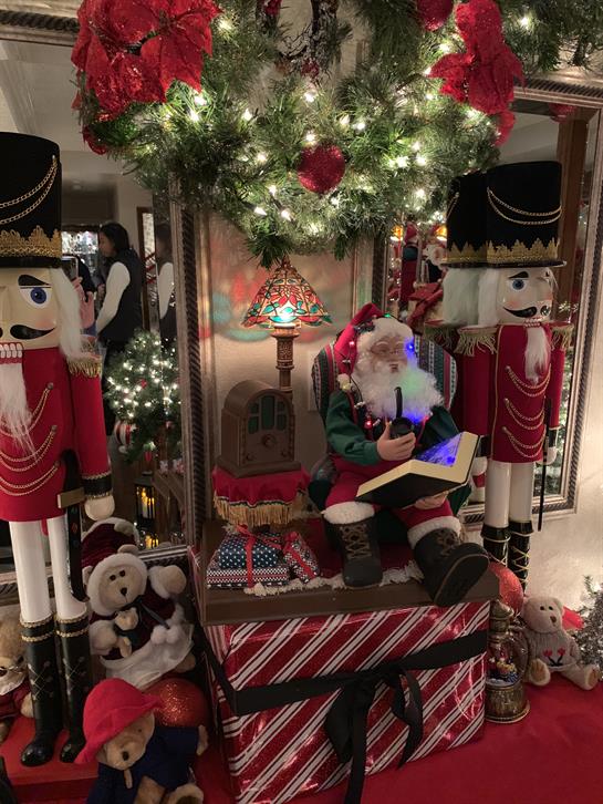 Santa sits by a beautifully decorated tree with nutcracker figures and plush toys surrounding him.