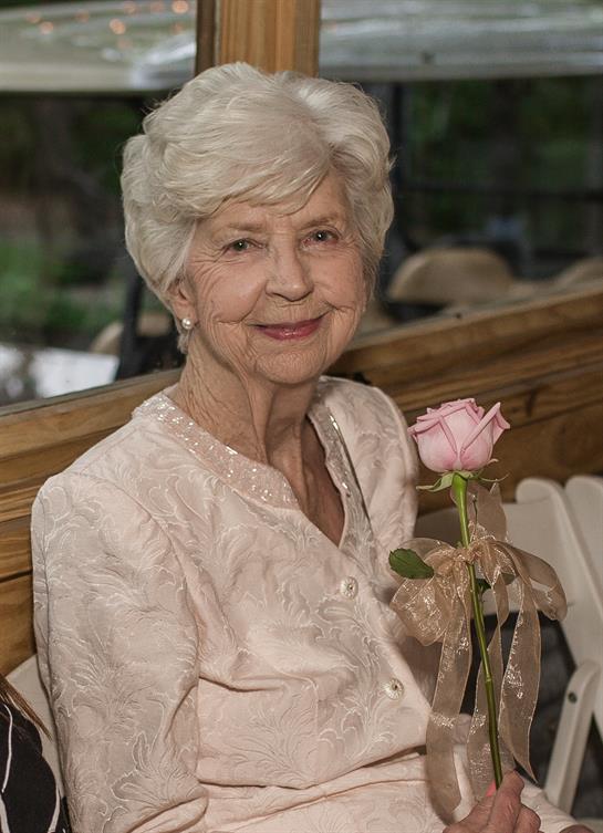 Senior woman smiles gently while seated on a bench, holding a delicate pink rose in a lush garden.