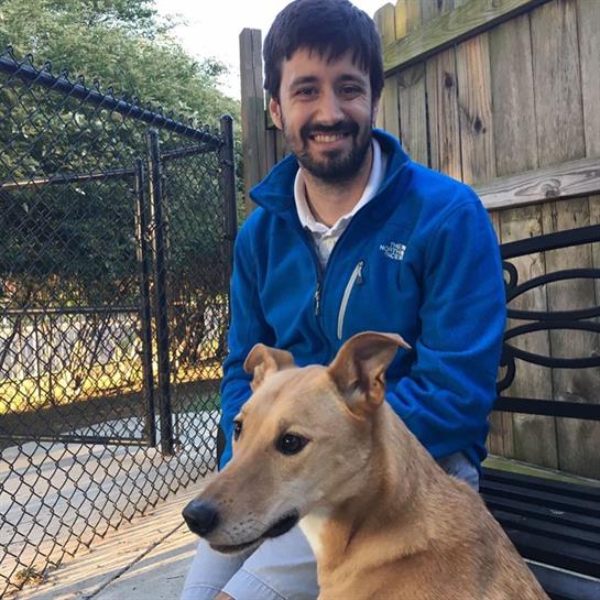 A man smiles while seated on a bench next to a friendly brown dog in a sunny backyard.