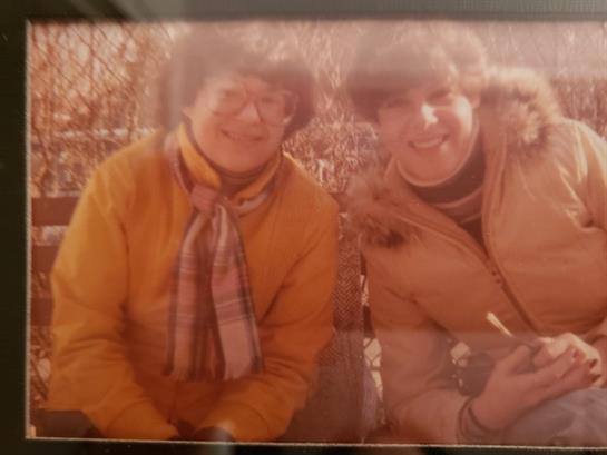 Two friends sit together outdoors in autumn, both dressed warmly and smiling brightly.