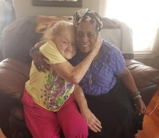 Two women embrace each other warmly while sitting on a couch in a sunny living room.