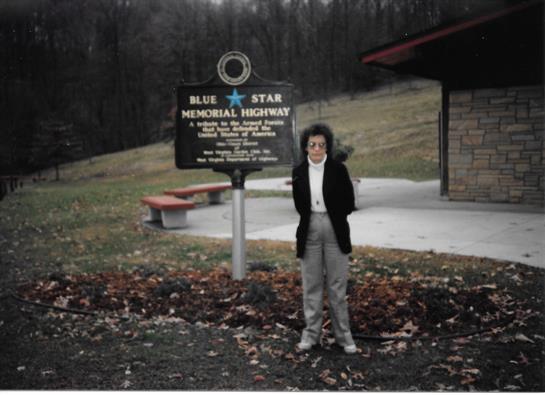 A person poses next to a historical marker for Blue Star Memorial Highway in a serene landscape.