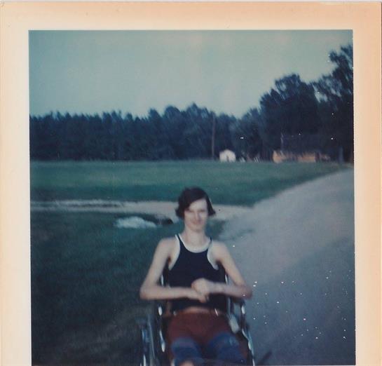 A young person in a wheelchair relaxes outdoors, surrounded by greenery and open space.