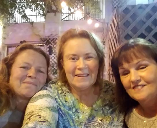 Three friends gather close together, smiling warmly for a selfie taken during a lively evening.