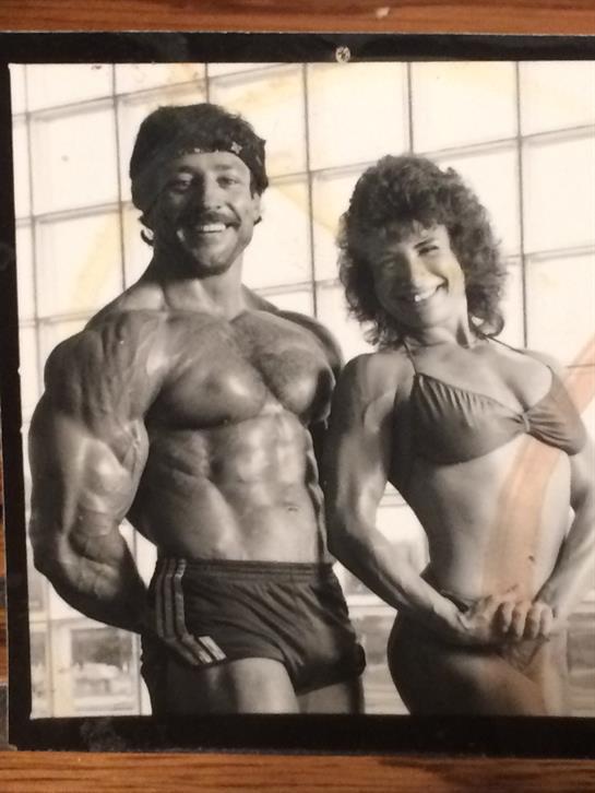 Two bodybuilders proudly showcase their physiques in a sunlit gym.