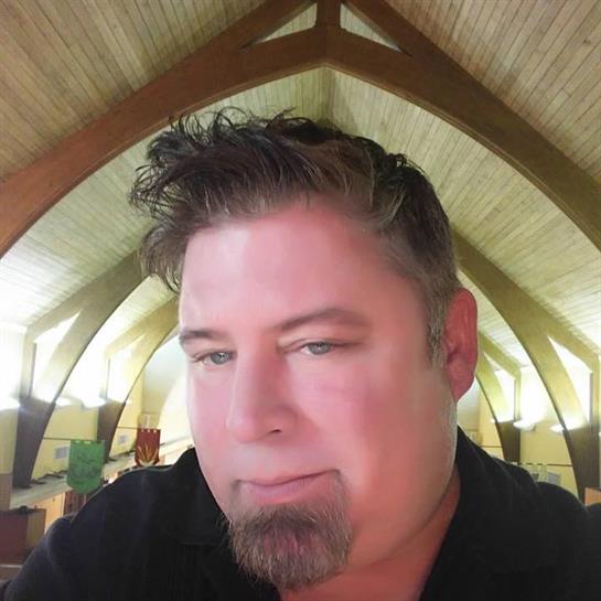 A man with a goatee sits calmly indoors, surrounded by a beautifully arched wooden ceiling.