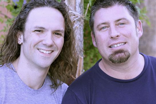 Two smiling men pose for a friendly picture in a natural setting surrounded by greenery.