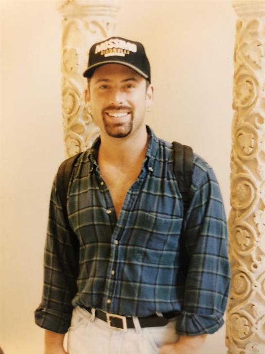 A man stands near decorative pillars, wearing a plaid shirt and cap, enjoying a sunny day.