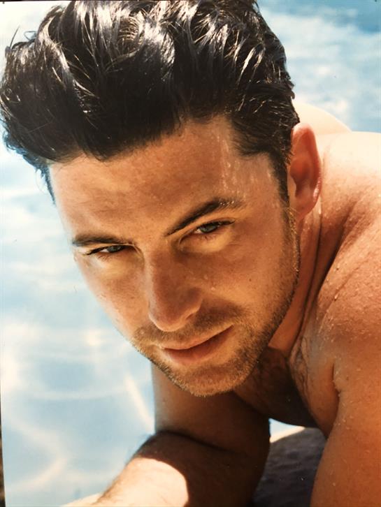 A male model is reclining on a surface near water, enjoying a sunny day outdoors.