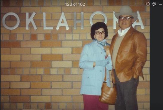Couple dressed in vintage clothing stands proudly near a brick wall marked Oklahoma during dusk.