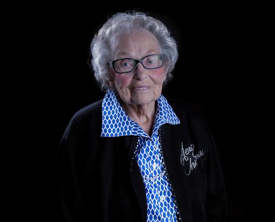 A senior woman with curly gray hair and glasses smiles in a blue patterned shirt and black jacket.