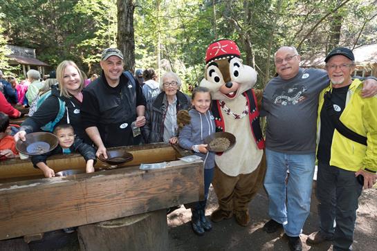 A family engages in gold panning while posing with a friendly cartoon character in a forest.