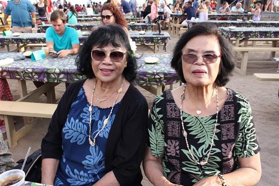 Two women wearing sunglasses pose together during a lively cultural festival in a crowded area.