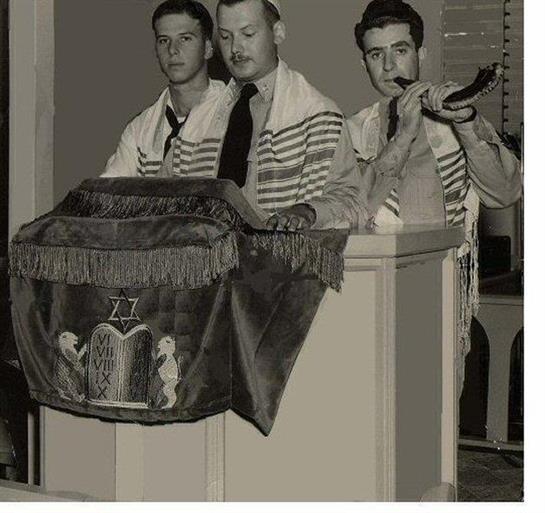Three men in traditional attire conduct a ritual in a synagogue, fostering a heartfelt atmosphere.