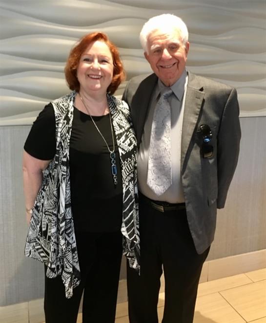 A couple stands side by side, smiling warmly in a contemporary indoor setting, dressed elegantly.