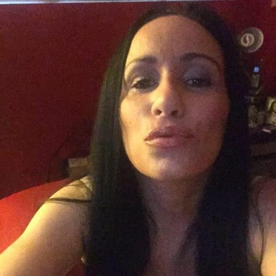 A woman with long black hair leans forward, blowing a kiss in a warmly lit red room setting.